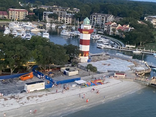 A colorful lighthouse near a marina, with boats docked and people on the beach, surrounded by trees and buildings.
