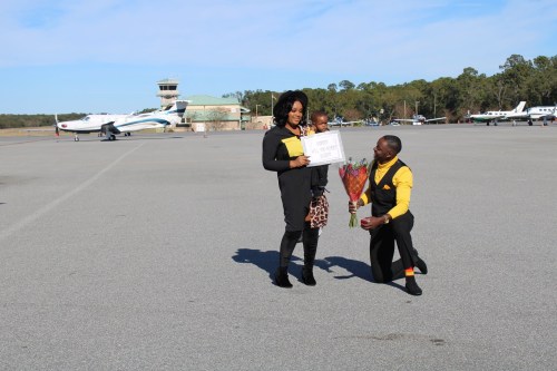 A man kneels with flowers while a woman holds a sign, both smiling with a child at an airport runway.