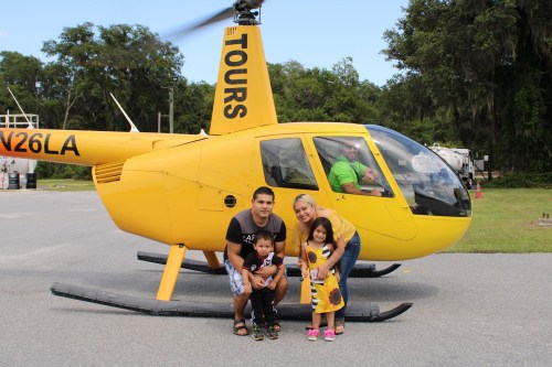 A family poses in front of a yellow helicopter, smiling, with a pilot visible inside the cockpit.