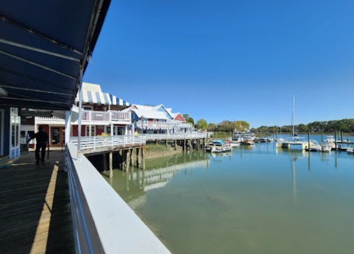 A sunny waterfront scene with colorful buildings, boats docked, and calm water reflecting the clear blue sky.