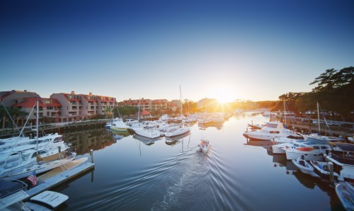 A serene marina at sunset, with boats docked and a small boat leaving a wake on the calm water.
