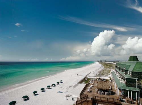 A serene beach scene with white sand, turquoise water, and a clear blue sky, featuring umbrellas and a coastal building.