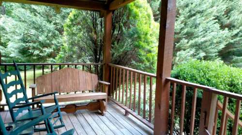A wooden porch with a bench and rocking chairs, surrounded by lush greenery and trees.