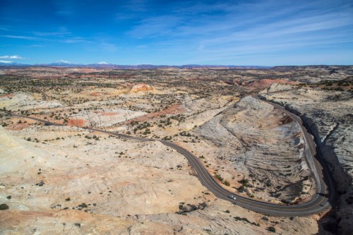 A winding road cuts through a vast, colorful desert landscape under a clear blue sky.