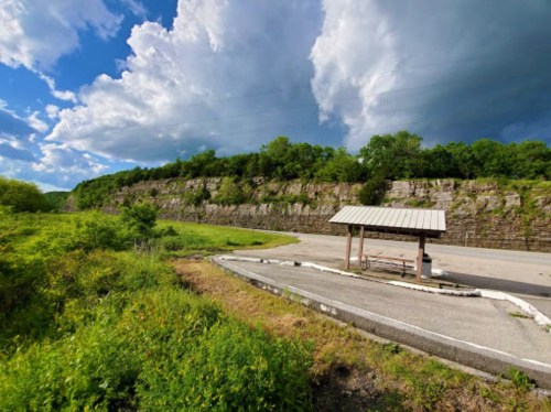 A scenic view of a parking area surrounded by lush greenery and a rocky cliff under a cloudy sky.
