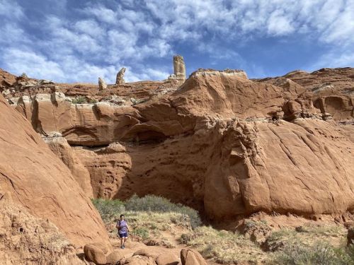 A person stands near red rock formations under a blue sky with scattered clouds.