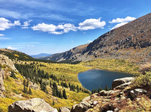 A scenic view of a valley with a lake, surrounded by mountains and trees under a blue sky with fluffy clouds.