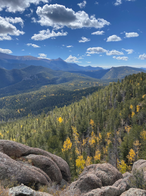 A scenic view of mountains and forests with autumn foliage under a blue sky with clouds.