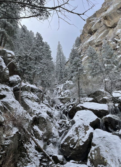 Snow-covered rocks and trees in a mountainous landscape, with a cloudy sky above.