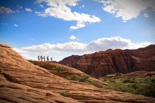 A group of hikers traverses rocky terrain under a blue sky with fluffy clouds and red rock formations in the background.