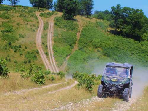 A rugged off-road vehicle drives along a dusty trail in a green, hilly landscape under a clear blue sky.