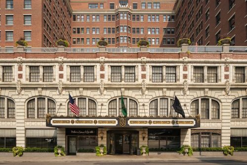 Historic hotel facade with ornate architecture, featuring flags and greenery, set against a tall brick building.