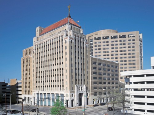 Historic building with ornate architecture, featuring a statue on top, surrounded by modern structures and a parking garage.