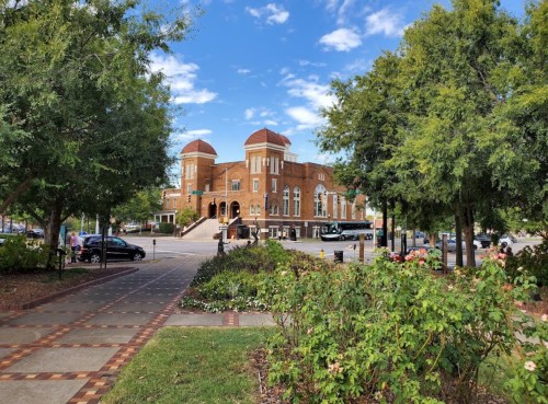 Historic brick building with domed roofs, surrounded by greenery and a pathway, under a blue sky with clouds.