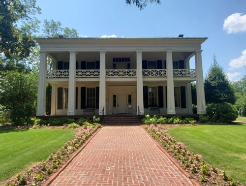 A historic two-story house with white columns, surrounded by greenery and a brick pathway lined with flowers.