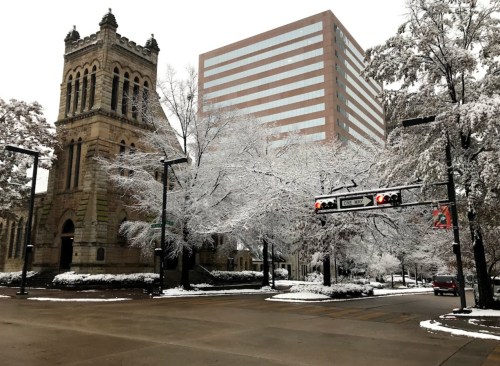 A snowy street scene featuring a historic building and a modern skyscraper, with trees covered in white snow.