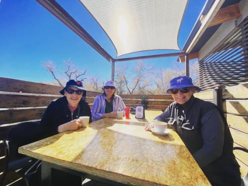 Three women wearing hats sit at a table outdoors, enjoying coffee under a shaded area with clear blue skies.