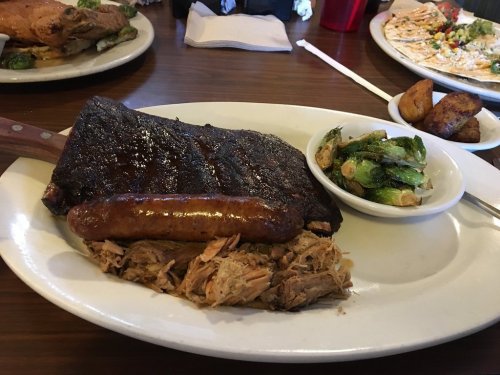 A plate featuring smoked ribs, sausage, pulled pork, and a side of Brussels sprouts and roasted potatoes.