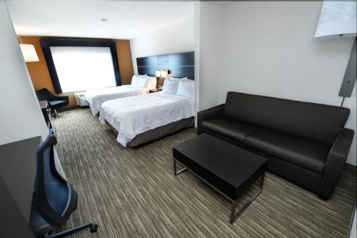 A hotel room featuring two beds, a sofa, a coffee table, and a desk, with modern decor and natural light.