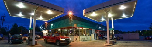 A gas station at dusk, with bright lights illuminating the building and cars parked nearby.