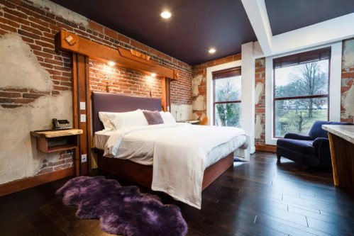 Cozy bedroom with exposed brick walls, a large bed, and a purple rug, featuring natural light from windows.