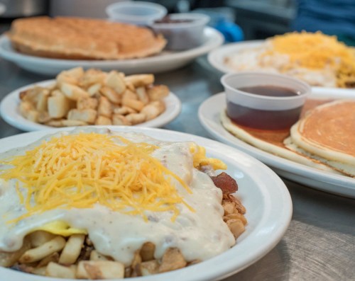 A plate of loaded breakfast hash topped with cheese, surrounded by pancakes, a waffle, and diced potatoes.