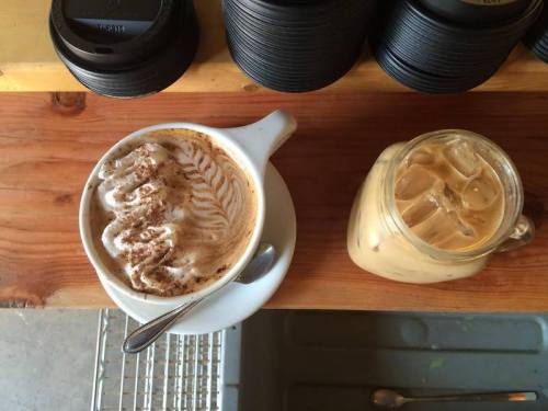 A cup of cappuccino with latte art and a glass of iced coffee on a wooden table.