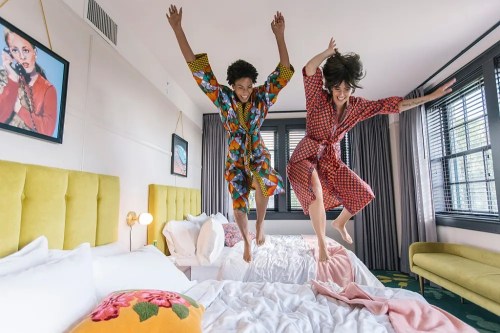 Two people in colorful robes joyfully jumping on a bed in a stylish hotel room.