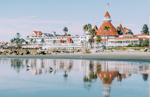 A beachfront view of a historic hotel with red roofs, palm trees, and reflections in the water.