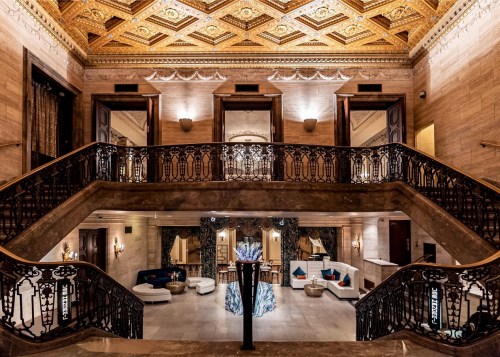 Elegant hotel lobby with ornate ceilings, grand staircase, and stylish seating areas. Warm lighting enhances the ambiance.