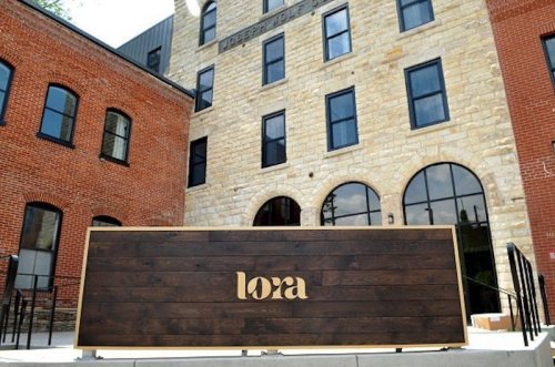 Exterior view of a building with a wooden sign reading "lora" in front of a stone and brick structure.