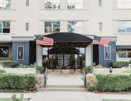Entrance of a building with an awning, flanked by American flags and surrounded by greenery.