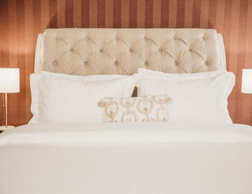 Cozy bed with a tufted headboard, white linens, and decorative pillows against a striped wall. Soft lighting from lamps.
