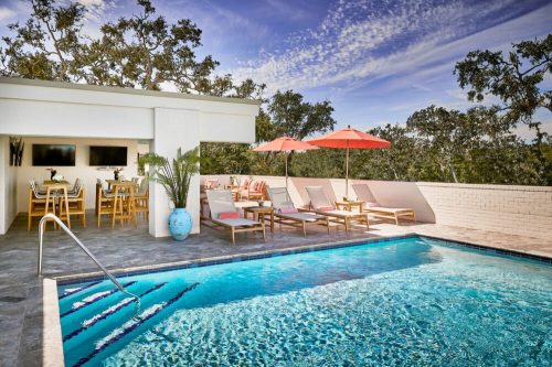 A rooftop pool area with lounge chairs, umbrellas, and dining space surrounded by greenery and a clear blue sky.