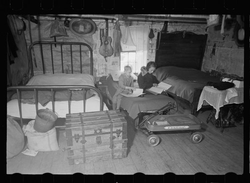 Two children sit on a bed in a rustic room, reading books beside a wagon and a trunk.