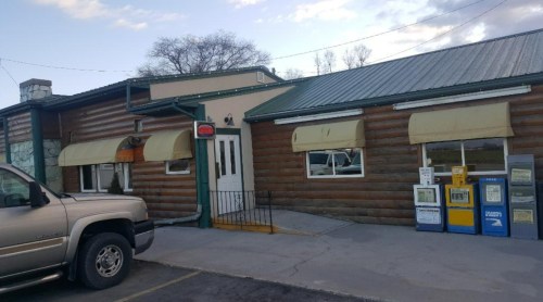 A rustic log cabin-style building with a green metal roof, featuring an open sign and newspaper boxes outside.