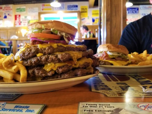 A towering cheeseburger with lettuce and tomato sits atop a plate of fries and a smaller burger in the background.