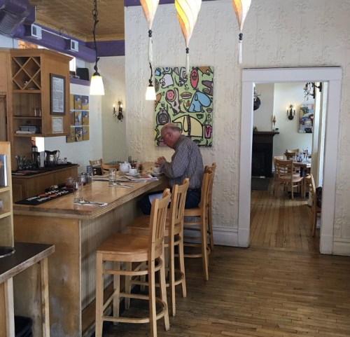 A cozy café interior with wooden floors, a man sitting at the bar, and colorful artwork on the walls.