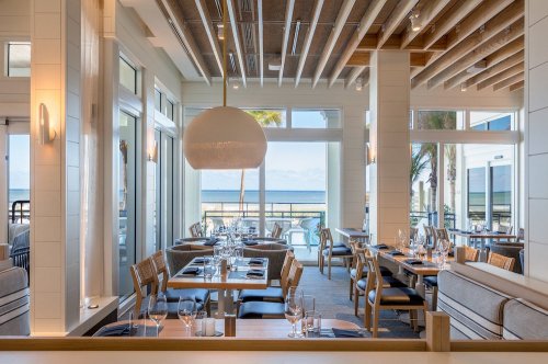 Bright, modern restaurant with ocean views, featuring wooden beams, elegant tables, and large windows.