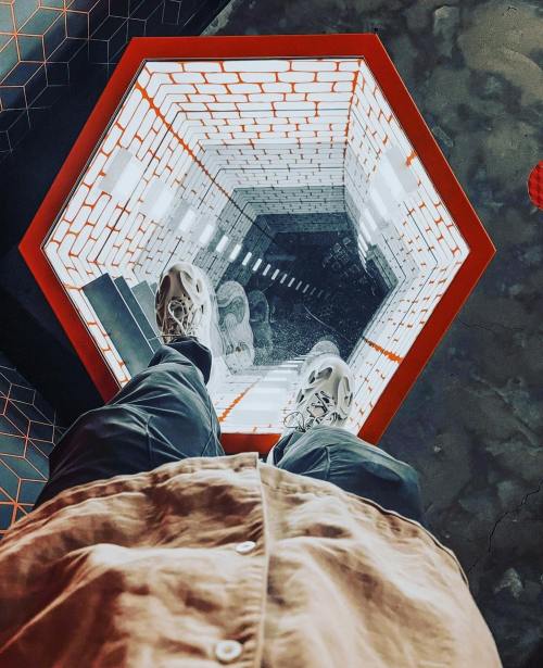 A person stands above a hexagonal mirror creating an illusion of a deep, endless tunnel.