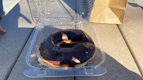 A chocolate-glazed donut in a clear plastic container, resting on a table with a brown paper bag nearby.