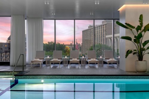 A serene indoor pool area with lounge chairs, large windows, and a sunset view of the city skyline.