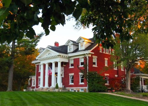 A large red brick house with white columns, surrounded by trees and a well-manicured lawn, in warm evening light.
