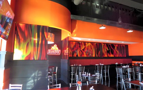 Interior of a modern restaurant with orange and black decor, featuring flame-themed wall art and metal chairs.