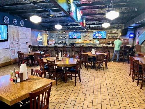 A spacious restaurant interior with wooden tables, chairs, a bar, and TVs displaying sports. A person stands at the bar.