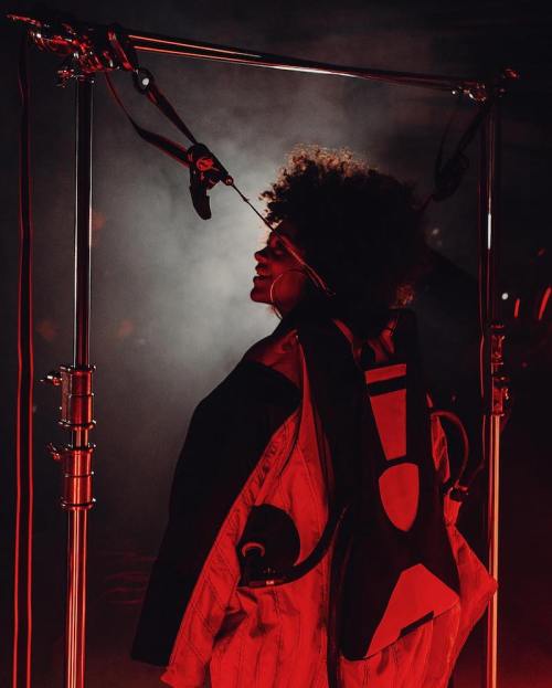 A person with an afro hairstyle stands in a dramatic red-lit setting, surrounded by equipment and smoke.
