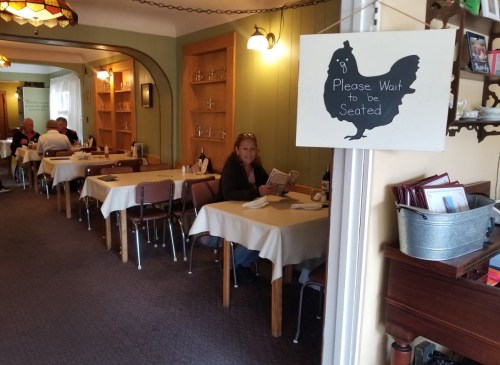 A cozy restaurant interior with tables set for dining and a sign that says "Please Wait to be Seated."