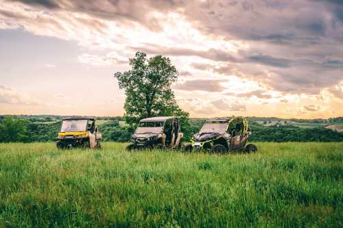 Three off-road vehicles parked in a grassy field under a cloudy sky, with rolling hills in the background.