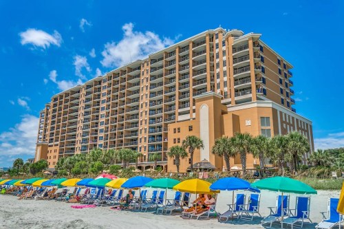 A beachfront resort with colorful umbrellas and lounge chairs in the foreground, under a bright blue sky.