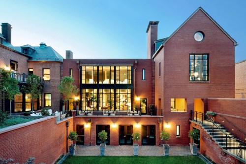 A modern brick building with large glass windows, surrounded by greenery and outdoor seating, illuminated at dusk.